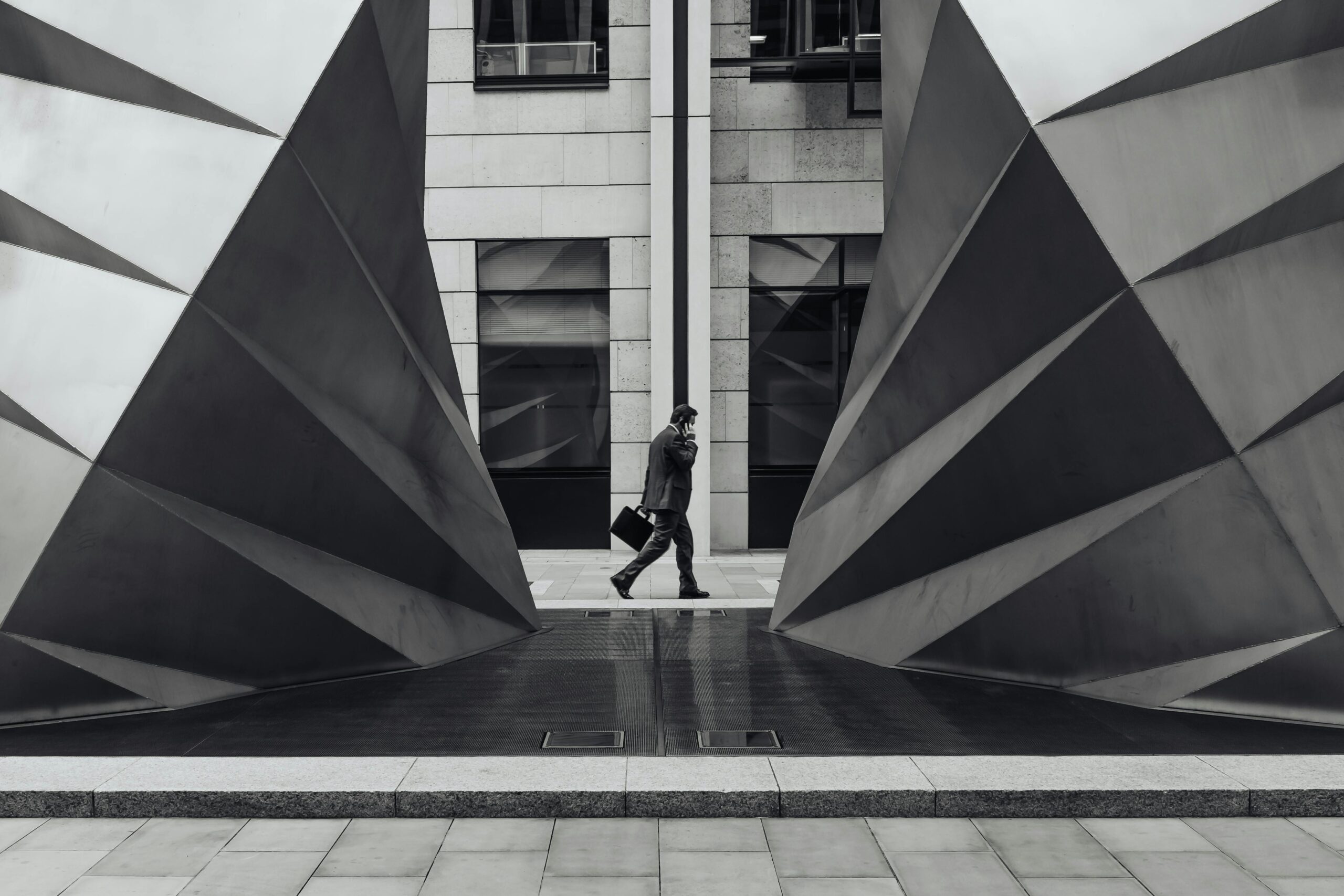 Monochrome photo of a businessman walking between geometric sculptures in a city setting.